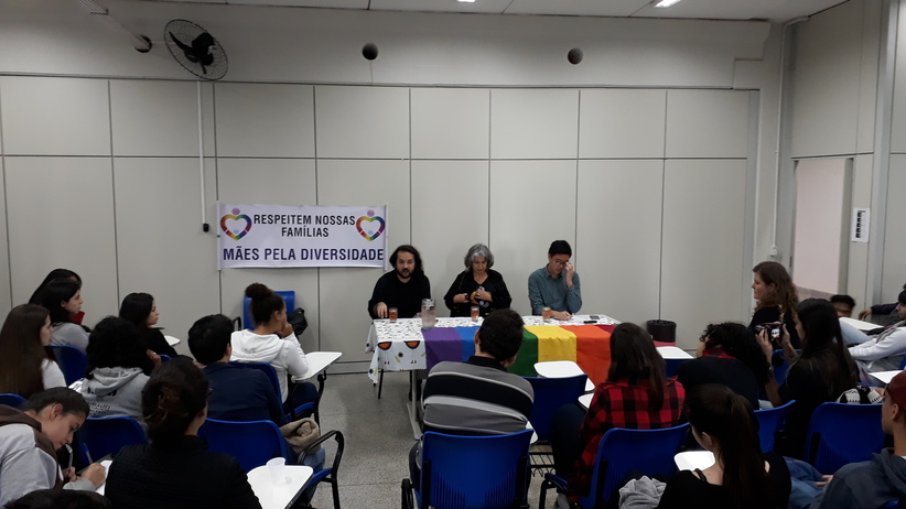 Palestrantes sentados frente à uma mesa com bandeira do movimento LGBT fazendo apresentação no evento sobre combata à homofobia no ambiente escolar. Platéia de servidores e estudantes fazendo perguntas