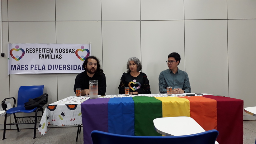 Palestrantes sentados frente à uma mesa com bandeira do movimento LGBT fazendo apresentação no evento sobre combata à homofobia