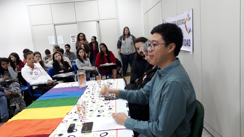 Palestrantes sentados frente à uma mesa com bandeira do movimento LGBT fazendo apresentação no evento sobre combata à homofobia