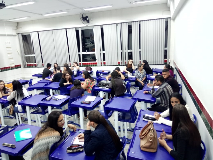 estudantes sentados em sala de aula com chão de cimento polido e paredes brancas com uma listra horizontal vermelha e outra verde, sentados em carteiras azuis interagindo uns com os outros