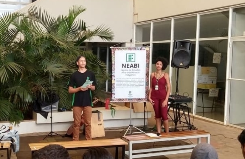 Um estudante e uma servidora de frente para uma plateia explicando sobre o Neabi. Ao findo um poster do NEabi em pedestal, instrumentos musicais e um jardim com palmeiras.