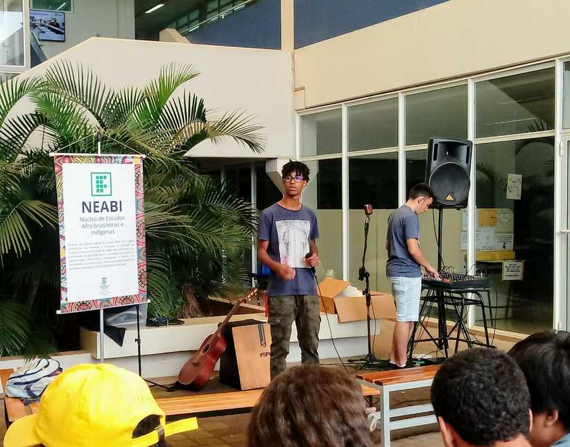 Estudante cantando com outro estudante manipulando a mesa de som. Ao lado esquerdo um poster do NEABI e um violão. Do lado direito uma caixa de som.