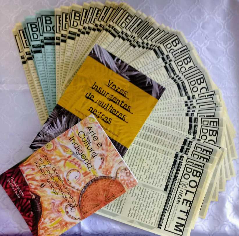 Foto com vários exemplares do boletim do Neabi dispostos em leque. Em cima do leque estão dois livros, um intitulado "Vozes insurgentes de mulheres negras" e outro intitulado "Arte e cultura indígena".