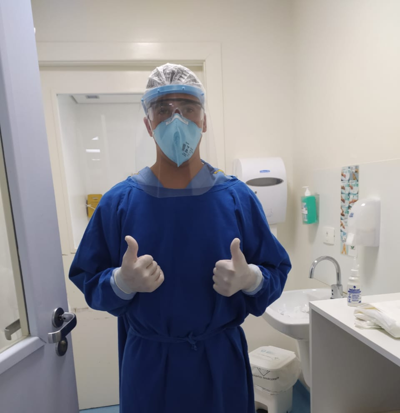 Enfermeiro em ambiente de trabalho usando roupa de proteção azul escuro, máscara de boca e nariz azul clara e protetor facial de acetato translúcido. Ele está em pé mostrando os polegares em posição de "jóia"