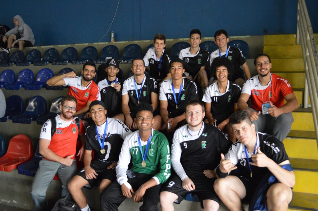 Fotografia colorida em ambiente interno que mostra integrantes da equipe masculina de Handebol do IFPR em uma arquibancada de ginásio de esportes. Os atletas vestem as medalhas e exibem o troféu de primeiro lugar.