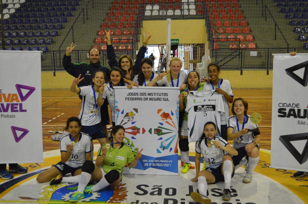 Fotografia colorida em ambiente interno que mostra a equipe feminina de Futsal do IFSul no centro de uma quadra de esportes. As atletas vestem as medalhas e exibem o troféu.