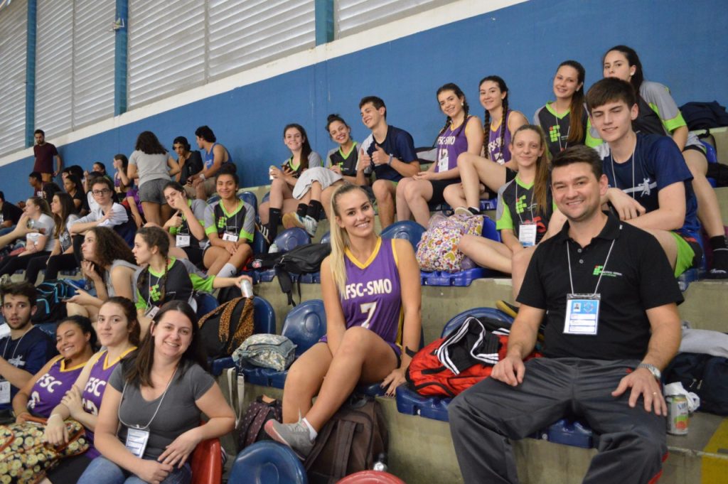Fotografia colorida em ambiente interno que mostra integrantes da delegação do IFSC nas arquibancadas de um ginásio de esportes.
