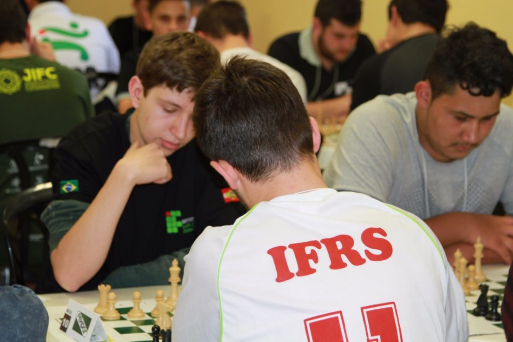 Fotografia colorida em ambiente interno que mostra, em primeiro plano, um atleta de costas. Ele observa um tabuleiro de Xadrez. Ao fundo, está o seu oponente. Ambos estão concentrados na partida.