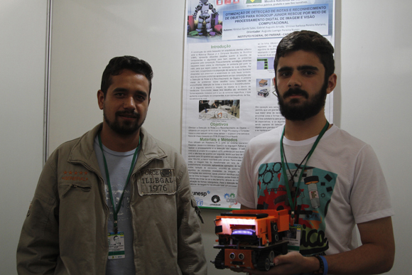 Professor e aluno posam para a foto segurando robô; ao fundo, banner traz explicações sobre o projeto
