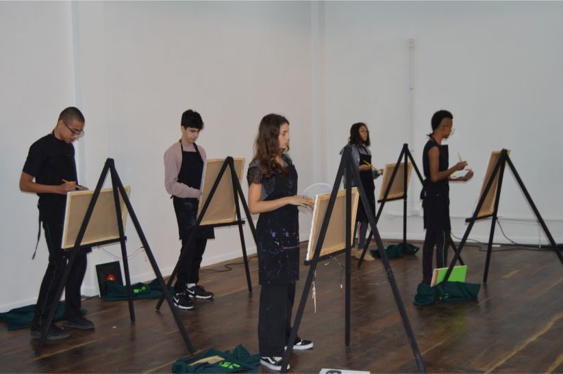 Foto de um ambiente fechado com estudantes pintando telas e um palco