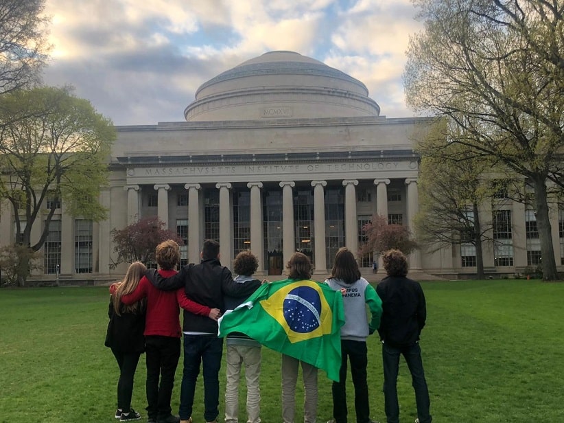 Fotografia colorida em ambiente externo. Em primeiro plano, estão os sete estudantes do Campus Capanema, eles estão lado a lado, abraçados, de costas. Um deles segura uma bandeira do Brasil, veste-a como se fosse uma capa. O grupo olha a fachada da universidade MIT, que está ao fundo.