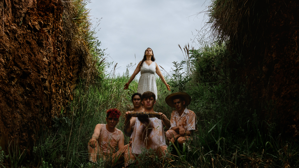 Foto dos atores do grupo de teatro. Duas mulheres e três homens. Vestidos de branco, sujos de poeira marrom. Os atores estão em um espaço cercado por vegetação alta dos dois lados.