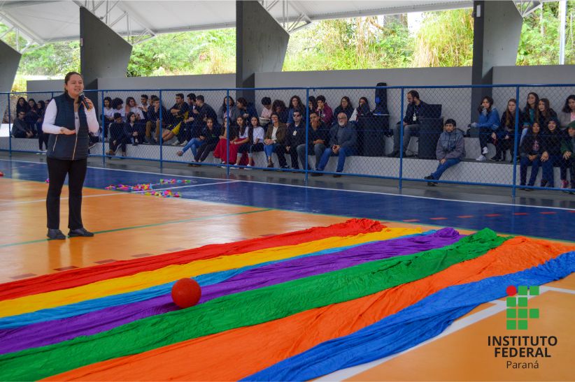 Professora de Educação Física faz uso do microfone para orientar atividades culturais. Na foto, publico composto por estudantes acompanha explicações. Uma bola vermelha e um pano de muitas cores estão no chão da quadra. 