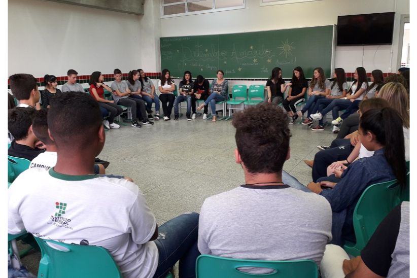 Fotografia colorida em ambiente interno. A fotografia mostra um grupo de estudantes, sentados em círculo em uma sala de aula do Campus Jaguariaíva. Ao fundo, em um quadro de cor verde, está escrito à mão, à giz, "Setembro Amarelo".