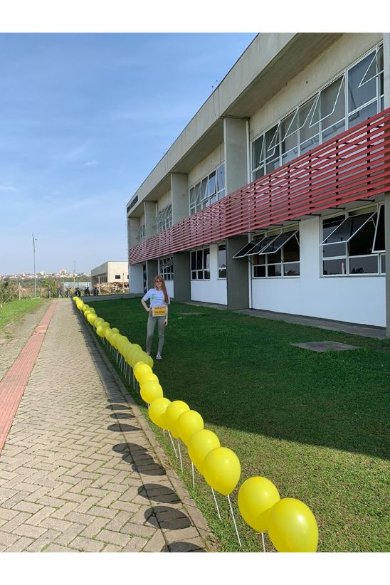 Fotografia colorida em ambiente esterno que mostra a lateral do prédio principal do Campus Pinhais. A calçada que acompanha a fachada está ladeada por balões amarelos.