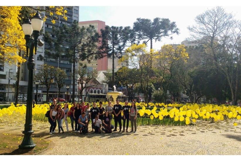 Imagem colorida em ambiente externo que mostra um grupo de estudantes na Praça Santos Andrade, em Curitiba. A praça está decorada com balões amarelos, que aparecem no fundo da imagem