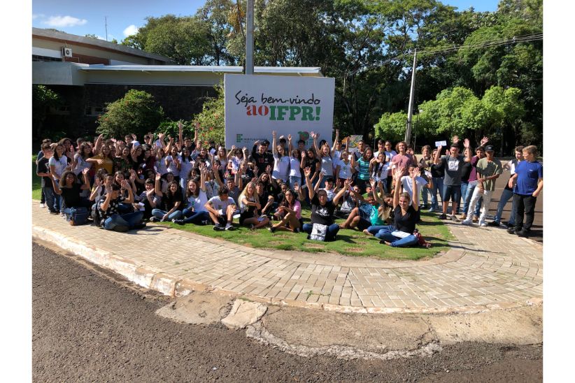 Fotografia colorida em ambiente externo que mostra um grupo de estudantes em frente ao Campus Foz do Iguaçu, junto a uma placa em que está escrita a frase "Bem-vindos ao IFPR".