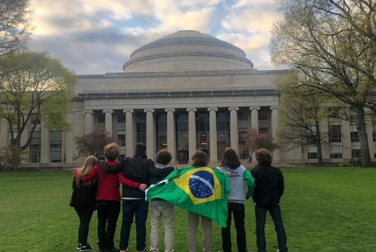 "Fachada de uma universidade. Há pessoas em pé com uma bandeira do brasil olhando para a estrutura"