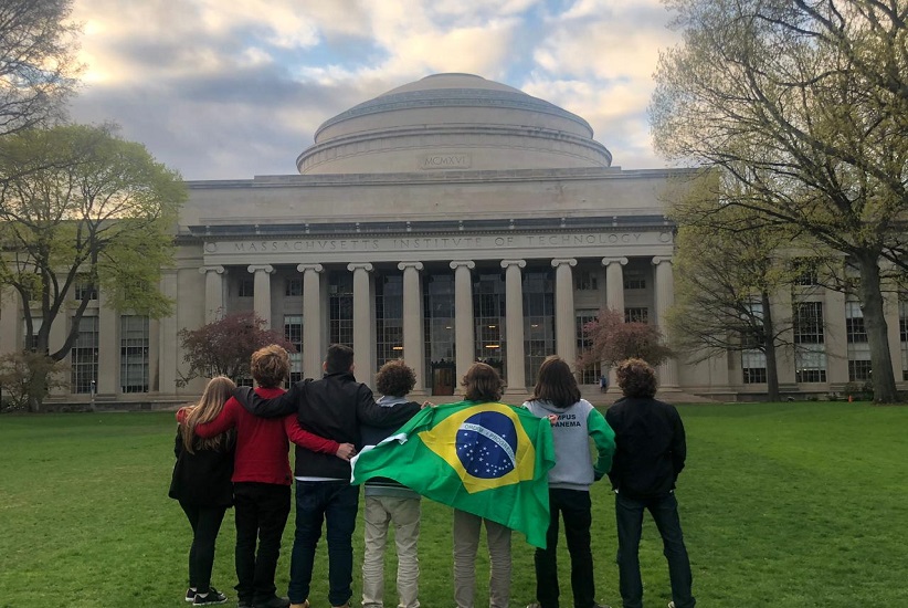 "Fachada de uma universidade. Há pessoas em pé com uma bandeira do brasil olhando para a estrutura"
