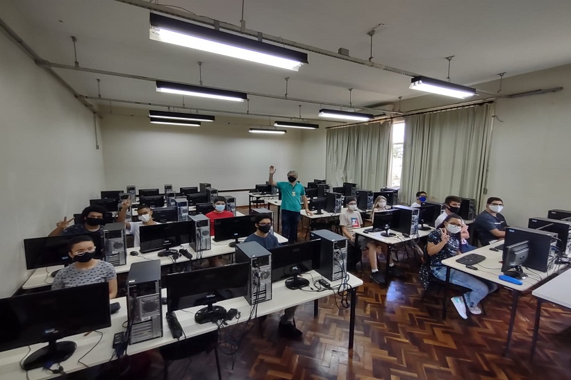 "Ambiente de sala de aula. Há estudantes sentados com computadores. No meio da sala há ujm homem em pé com a mão levantada"