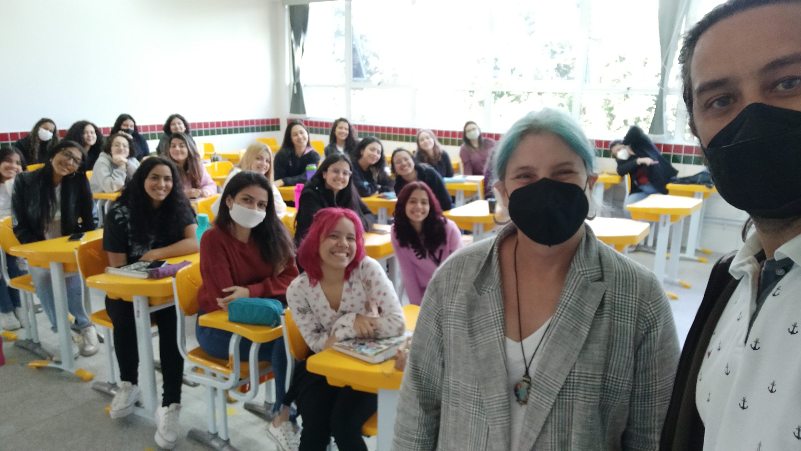 A imagem é uma foto, que mostra a pesquisadora ao lado do professor Leandro. Ao fundo, há uma sala de aula com cerca de 20 alunos, sorrindo para a foto.