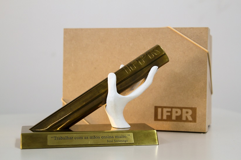 "Na imagem há uma caixa com a logo do IFPR. Na frente da caixa há a honraria Mão de Saramago"