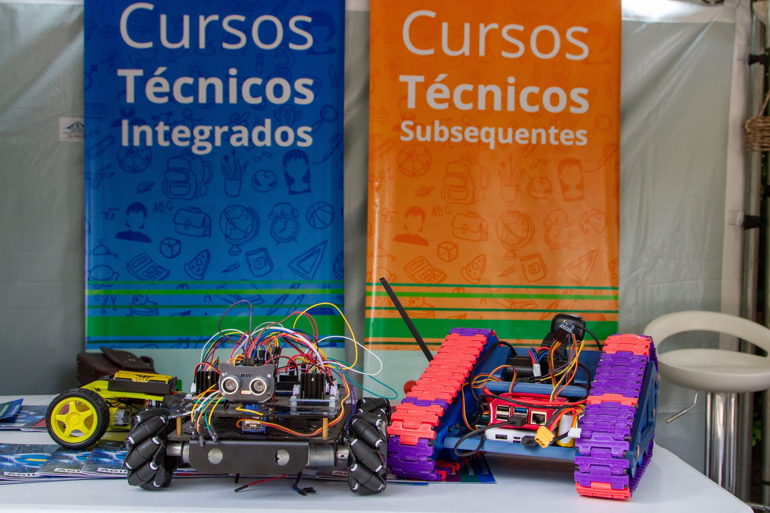 Fotografia em plano fechado com robôs à frente e dois banners coloridos atrás escrito: Cursos técnicos integrados e subsequentes