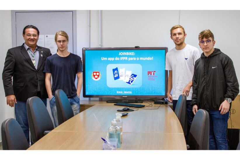 Imagem em ambiente interno que mostra, em primeiro plano, um grupo de quatro pessoas ao lado de uma tela de TV com informações sobre o projeto.