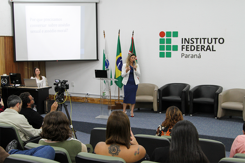 Servidores do IFPR sentados no auditório da Reitoria de frente para o palco, a Procuradora-Chefe do IFPR, Carliane de Oliveira Carvalho está no palco falando.